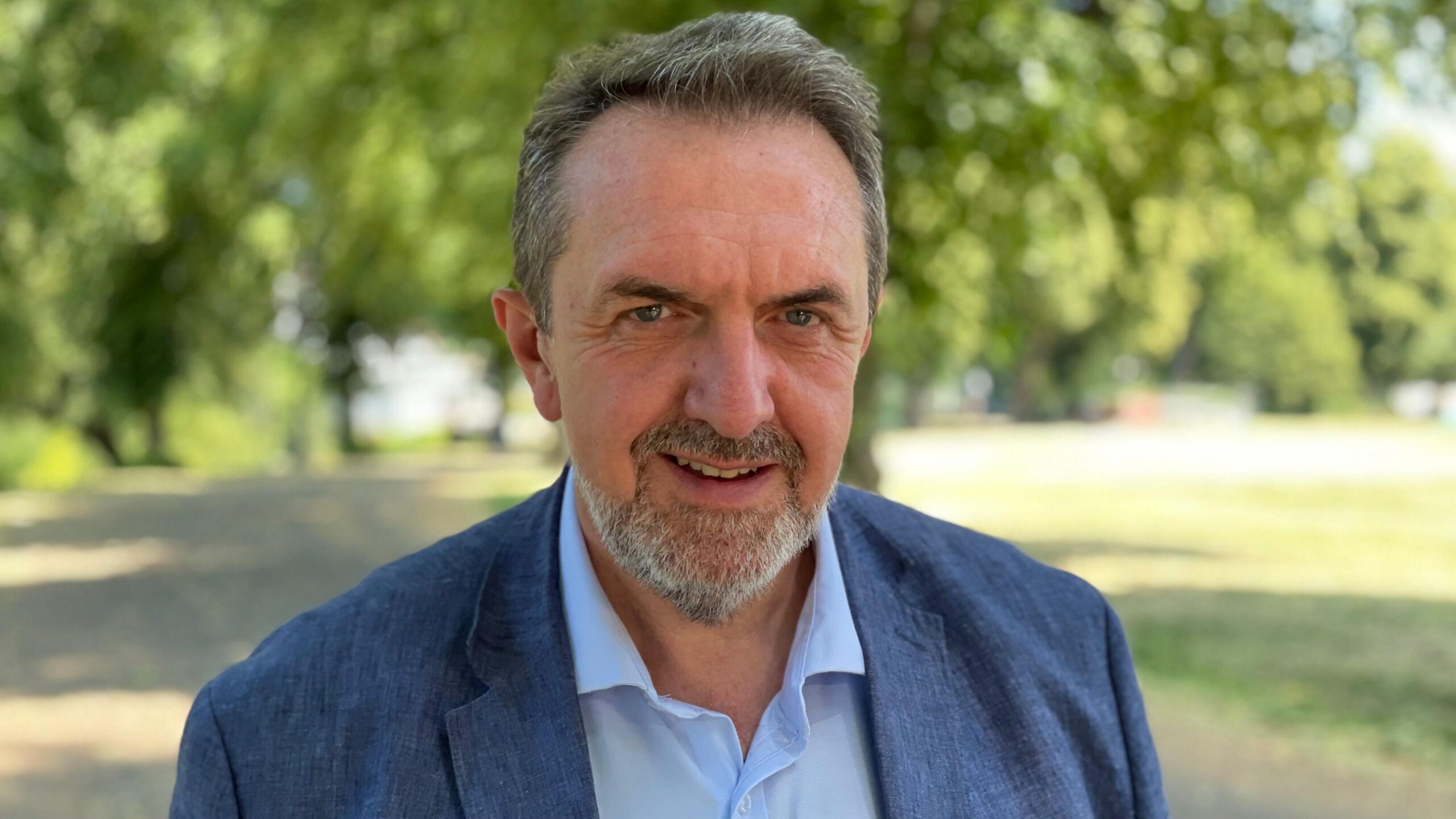 Matthew Green, looking directly down the camera. He is standing outside, with grass and trees behind him. Green is wearing a blue linen jacket and an open-necked blue shirt. He has dark hair, flecked with grey and a greying, neatly trimmed beard.
