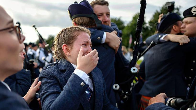 An emotional female piper, wearing a traditional outfit and with her hair in a French plait, is standing with one hand covering her mouth and part of her face. She is surrounded by other pipers who are hugging each other