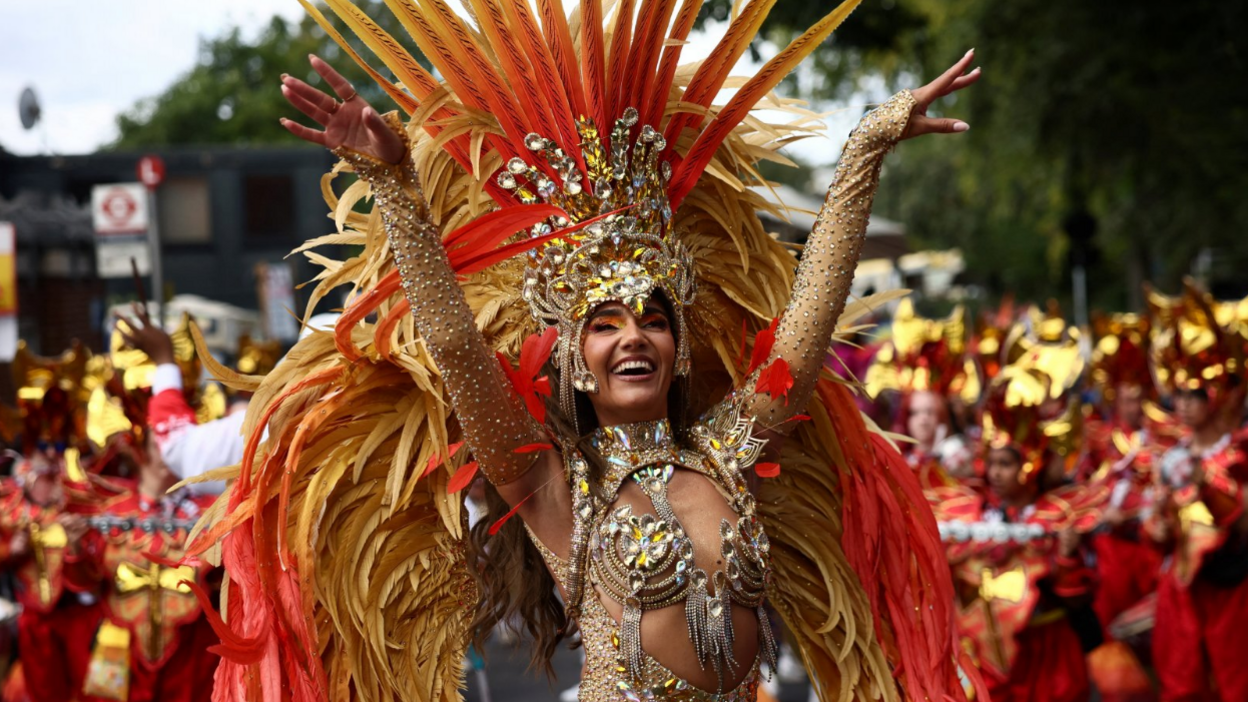 A woman in a gold and red beaded costume with feathers smiles and raises her arms into the air.
