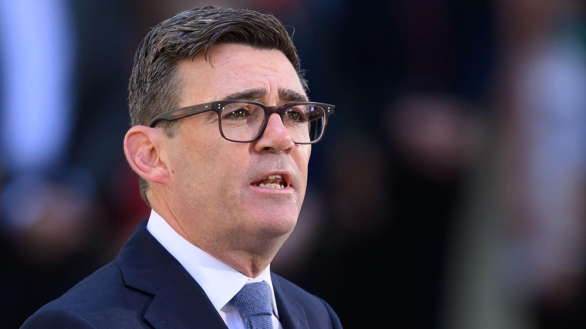 A close up shot shows Andy Burnham speaking outside Downing Street, wearing glasses and a dark suit and tie, with a bokeh background.