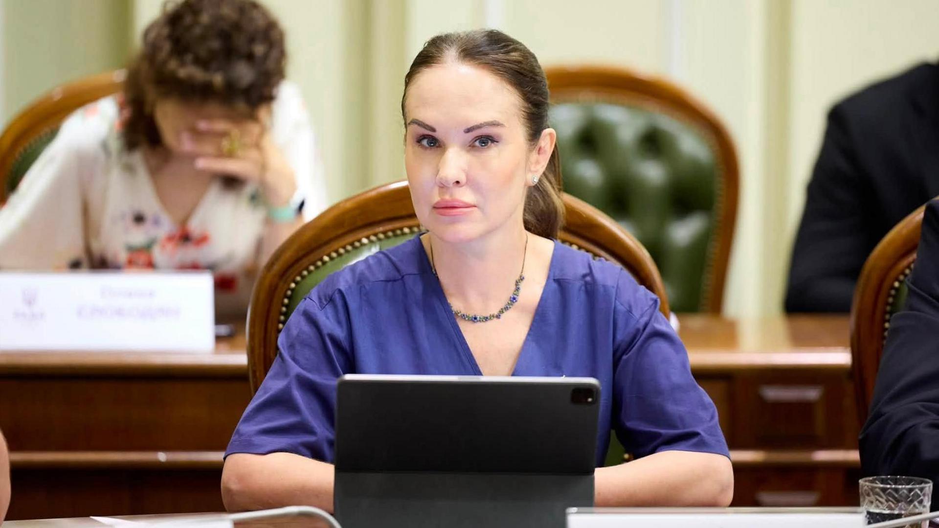A woman sits behind a laptop, with her hair up, wearing a blue blouse