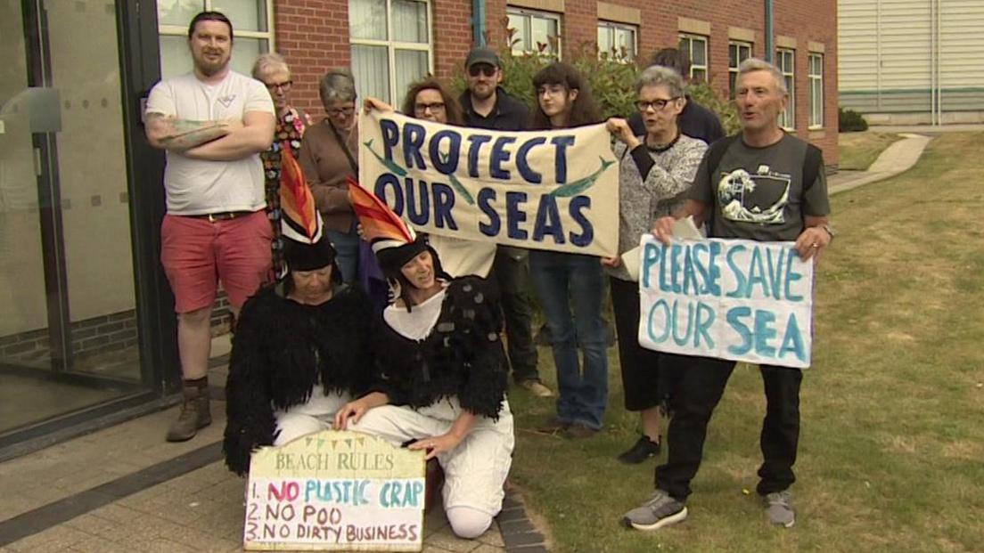 A group of campaigners outside the Port of Tyne Maritime House office, waiting to meet the firm's chief executive Matt Beeton. Two are knelt on the floor in puffin costumes. Others stand behind holding banners. One reads: "Protect our seas." Another reads: "Please save our sea." A third placcard reads: "Beach rules: No plastic crap, no poo, no dirty business."