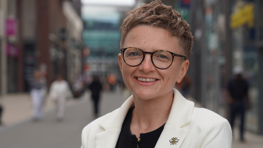 Bev Craig has short red hair, and is wearing glasses and a white jacket over a black blouse. She wears a Manchester Bee badge on her jacket lapel.