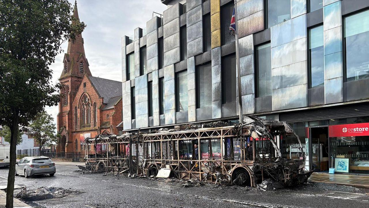 A Glider bus was set on fire in east Belfast. A burned out bus is in the foreground on a road which has visible marks of a fire. The bus is beside a large pale coloured building with lots of windows, and bearing scorch marks. To the rear of the picture is a red building, with a tall spire.