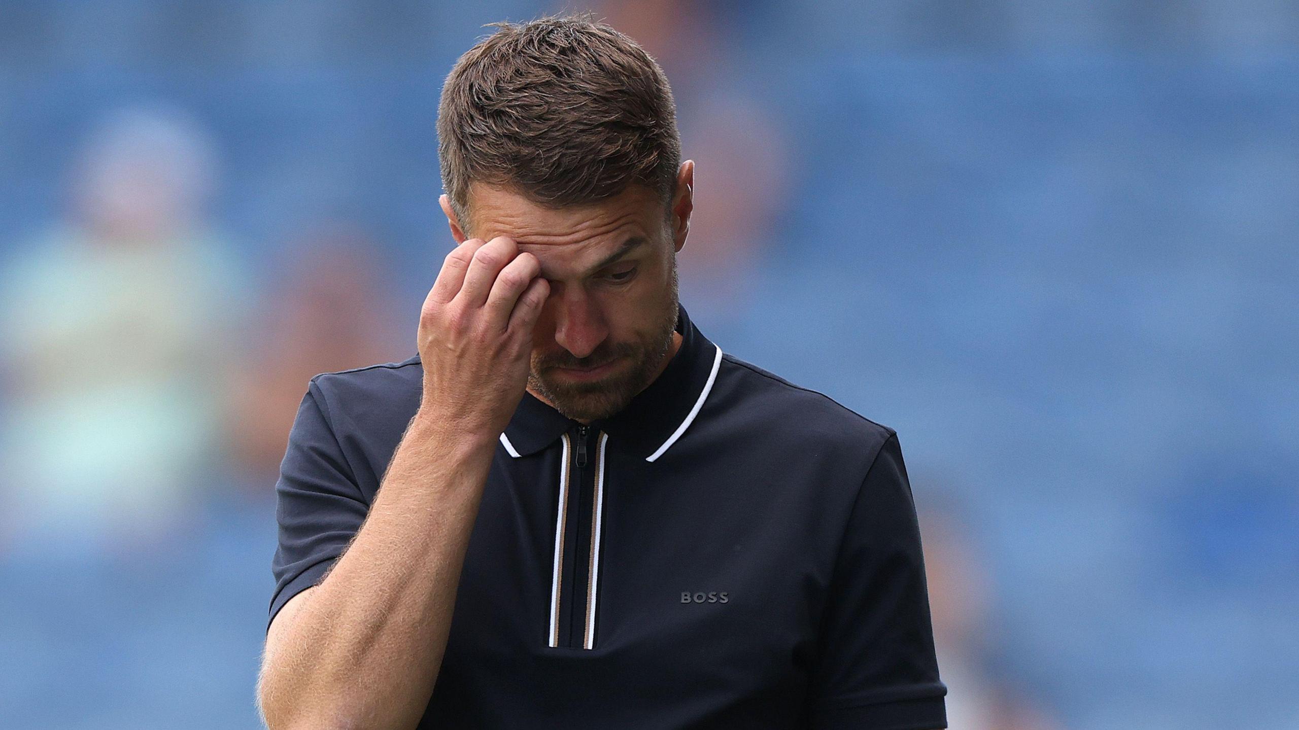 Aaron Ramsey, wearing a black Boss shirt with white trim, scratches his forehead during Oxford United's pre-season game against Ipswich Town