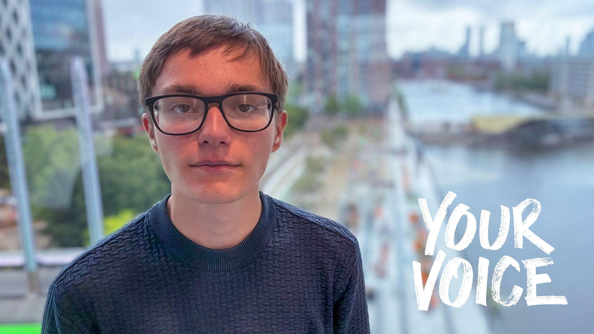 A young man wearing glasses looks into the camera with a blurred Salford docks in the background
