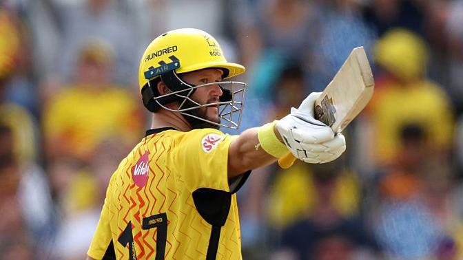 Ben Duckett (photographed from behind), wearing a yellow top and helmet, points his bat in his extended right arm in celebration