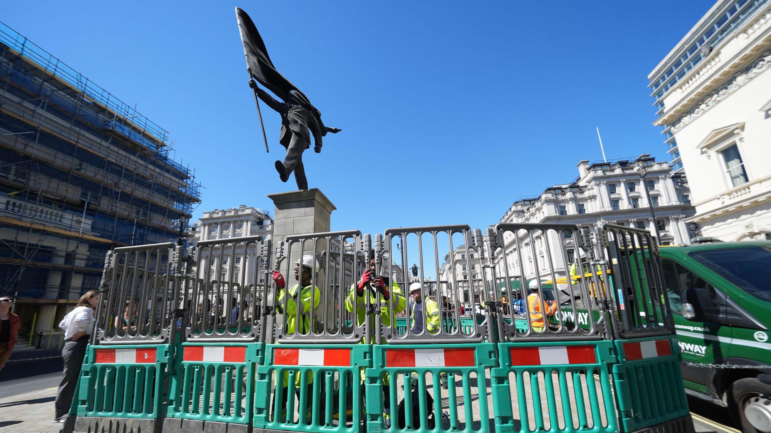 Plastic safety barriers being erected by workmen in high-vis jackets, in front of Banksy's statue of a man carrying a flag.