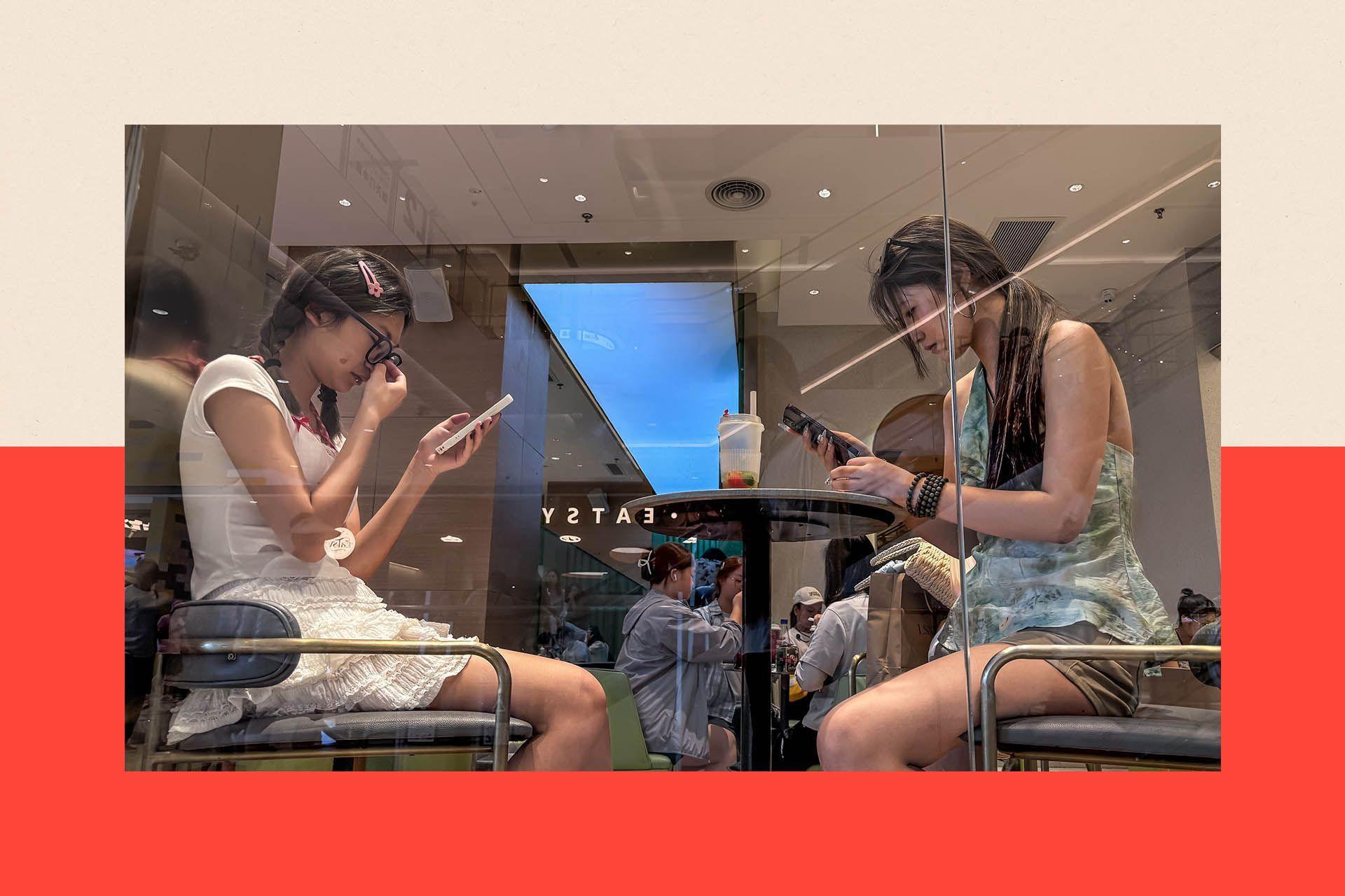 Two young women sit at a table on their phones