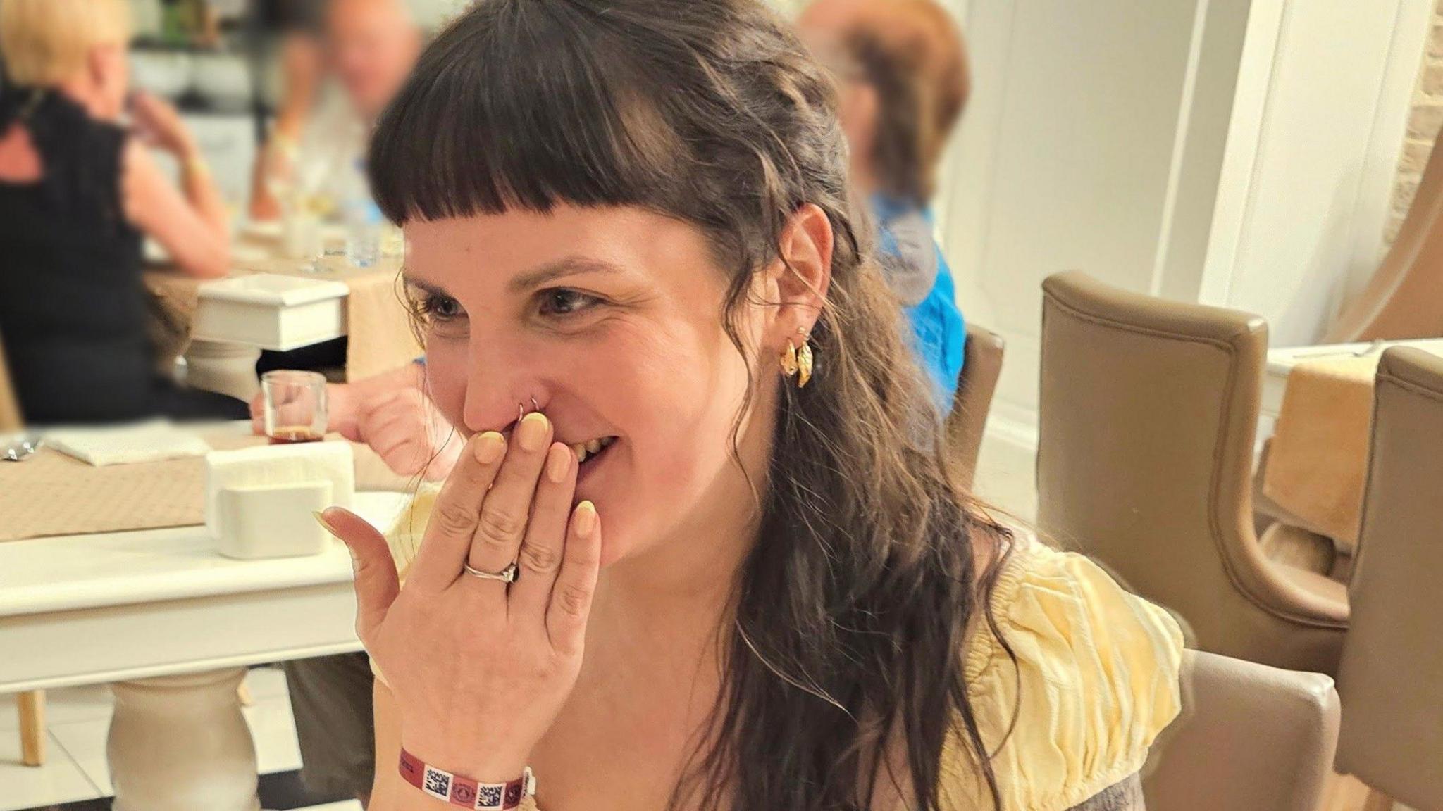 A woman with long brown hair and a yellow top sitting in a restaurant. She is laughing and is covering her mouth with her hand.