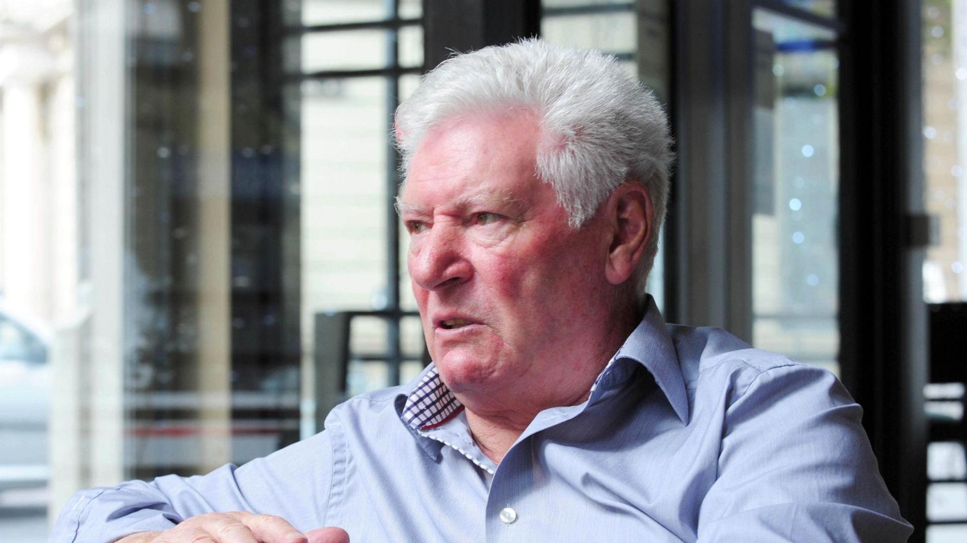 A man wears a blue shirt with the top button undone. He looks to his right, with his right arm leaning over on the top of a green and brown striped sofa. He has short white hair and brown eyes.