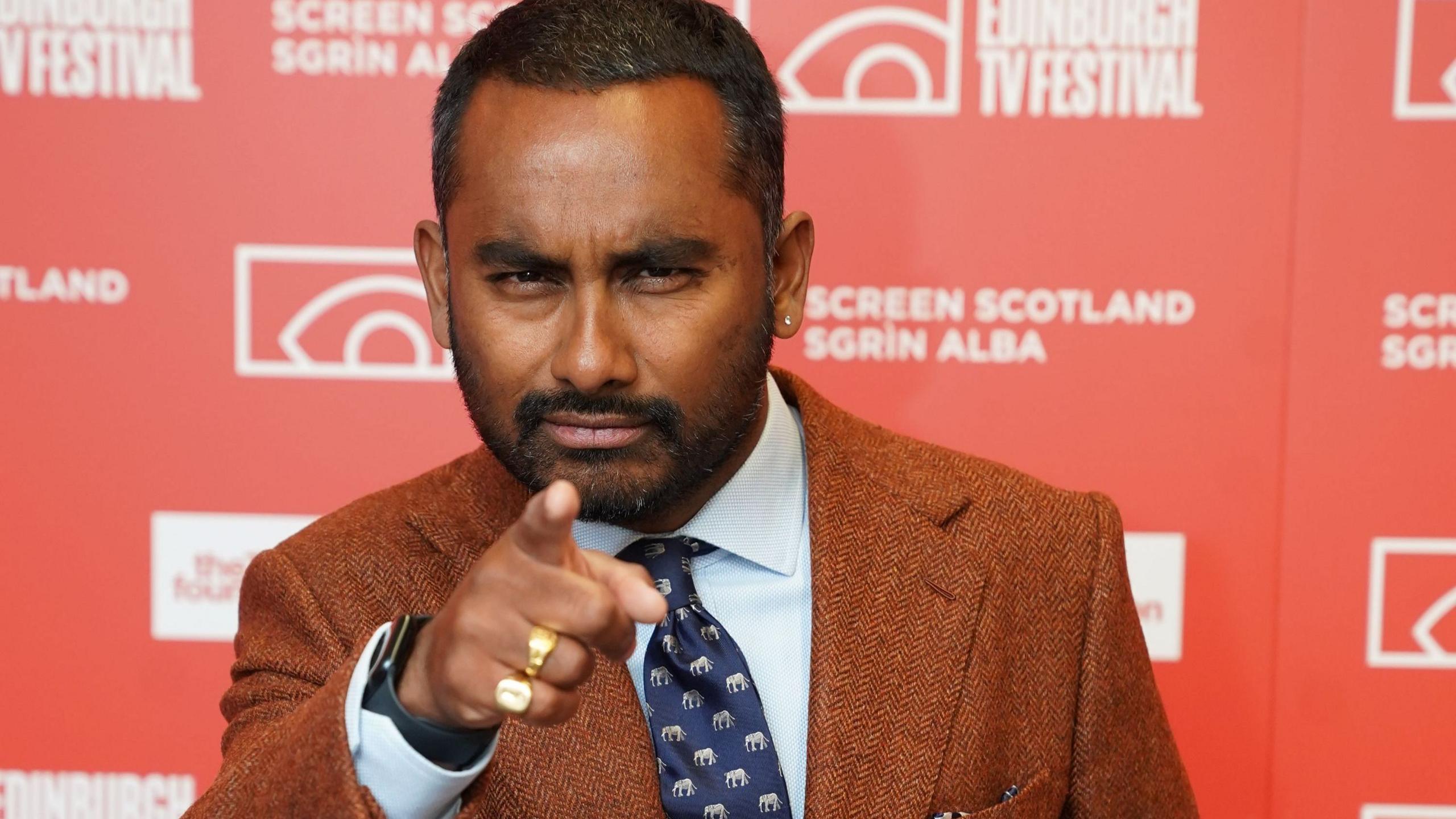 Presenter Amol Rajan, in a brown tweed jacket, pale blue shirt and navy tie, makes a playful face as he points at the camera at his lecture. The red backdrop carries all the TV festival's logos and sponsors.