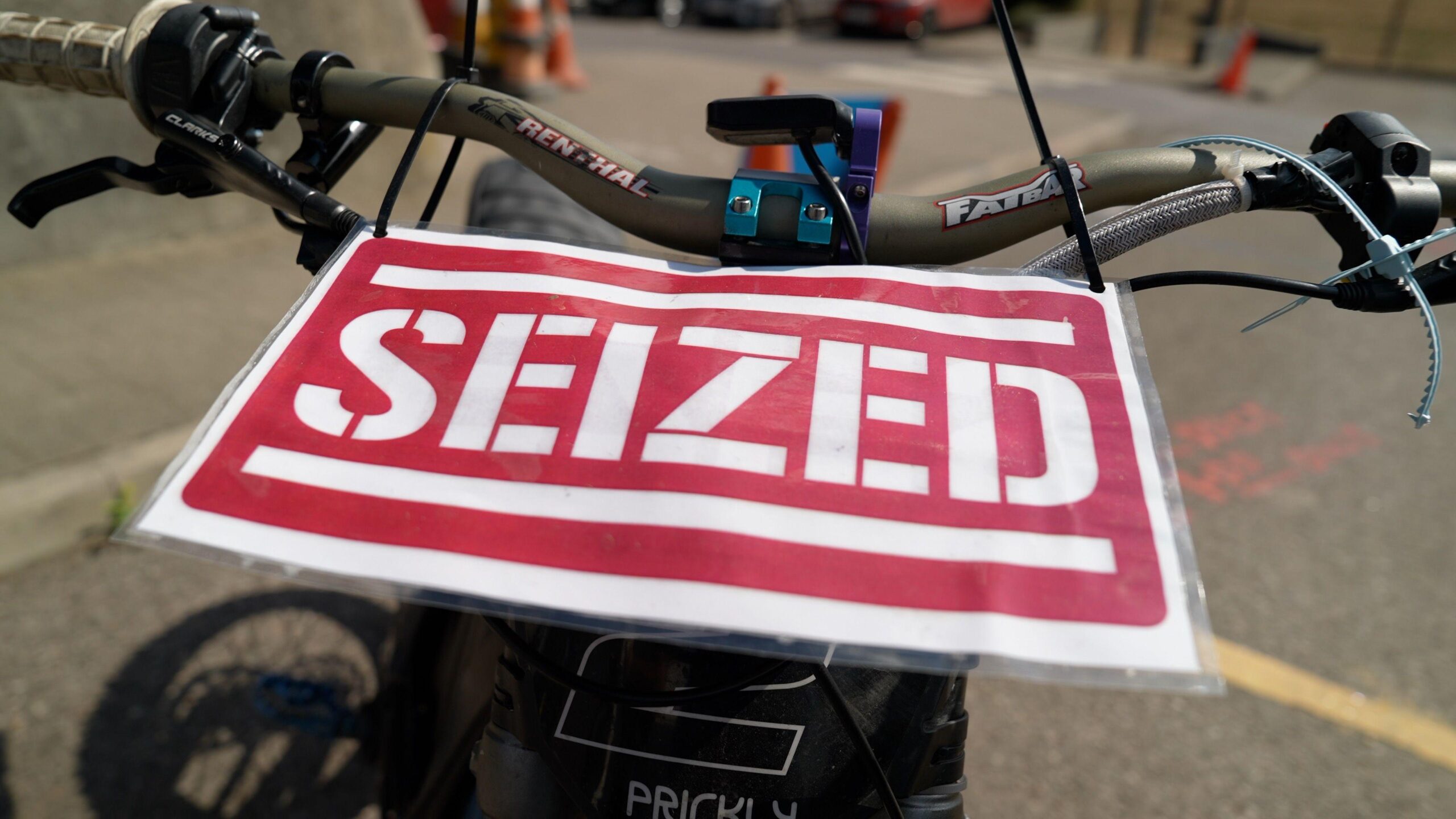 A red laminated paper sign which reads "seized" in capital letters is attached with cable ties to the handlebars of a bike. In the background there is concrete but not much of the bike is visible other than the brakes and wires and cables.