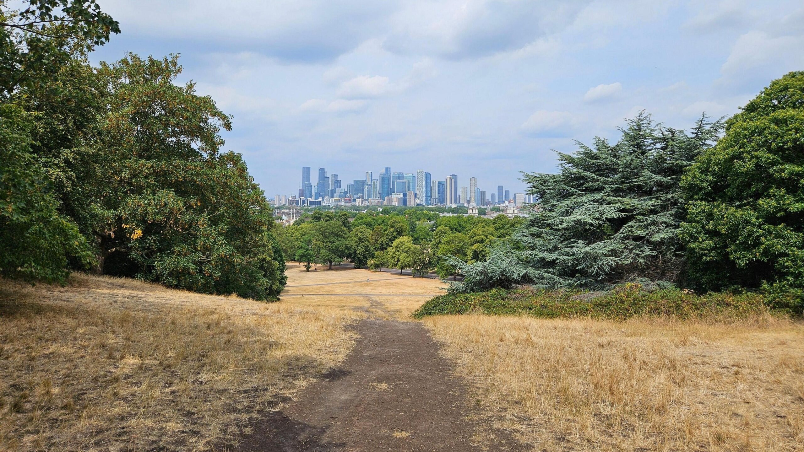 Dry ground and grass in a London park with the city skyline in the distance beneath a partly cloudy sky