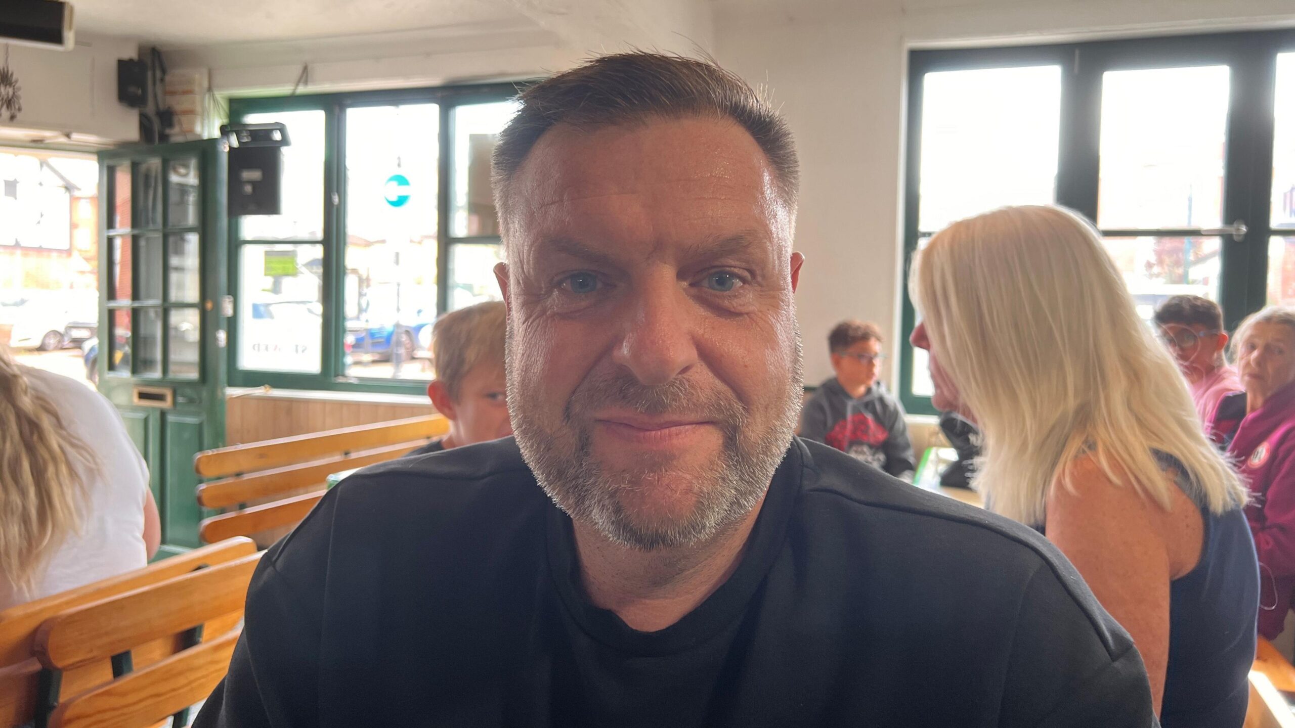 A man in his late 40s with a stubble beard and slicked-back hair, wearing a black T-shirt, looks directly into the can