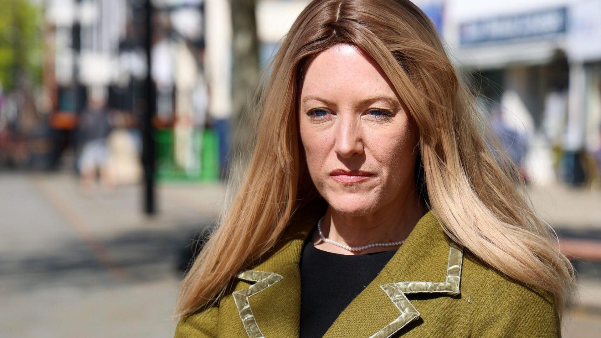 A head and shoulders photo of a woman with long blonde straight hair, wearing a green jacket. A blurred high street can be seen in the background.