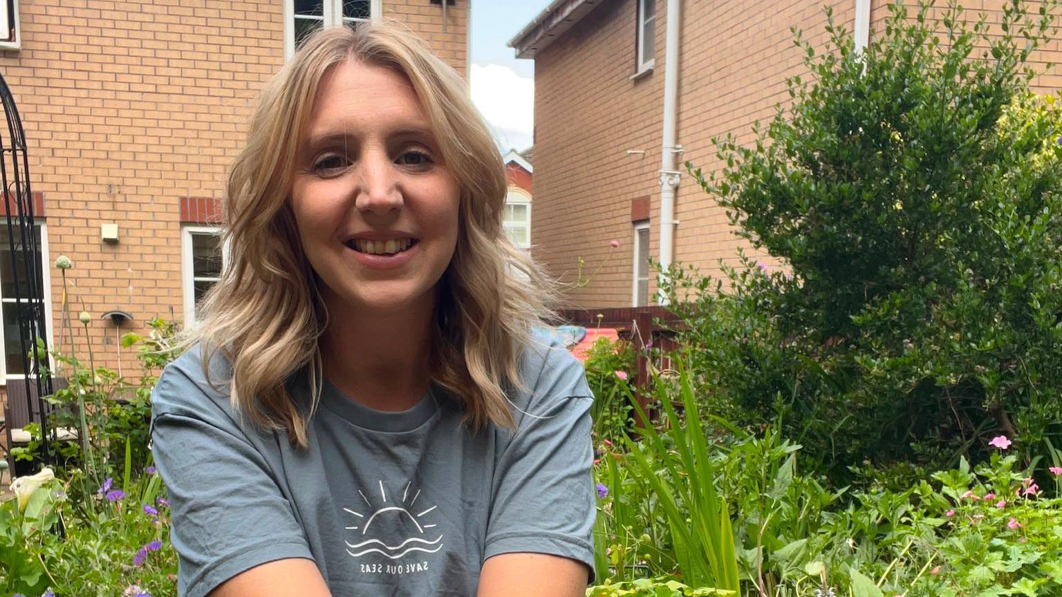 Rachel is sat in her garden, smiling. She has blonde hair and is wearing a grey/blue t-shirt.