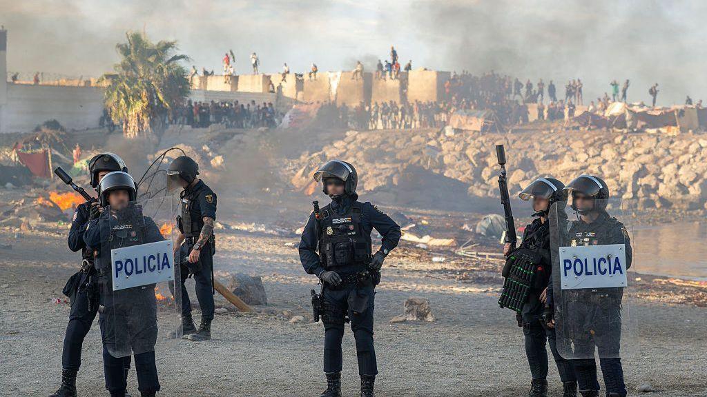 Spanish armed police with riot shields on Trampolin beach, Ceuta, as anti-migrant protesters set fire to a migrant camp there. Their faces are blurred. Smoke can be seen rising in the air.