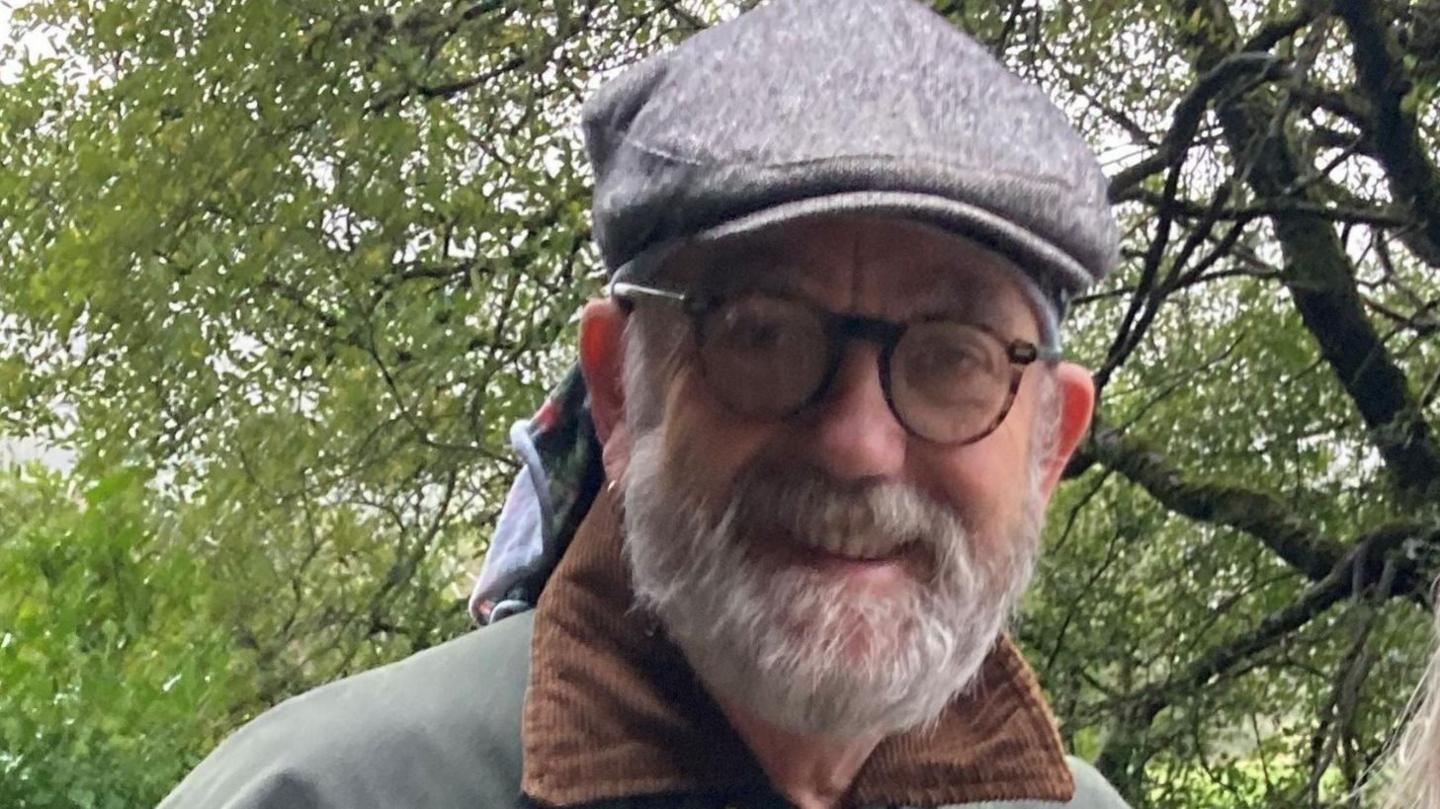 A man with a beard and glasses, wearing a grey flatcap