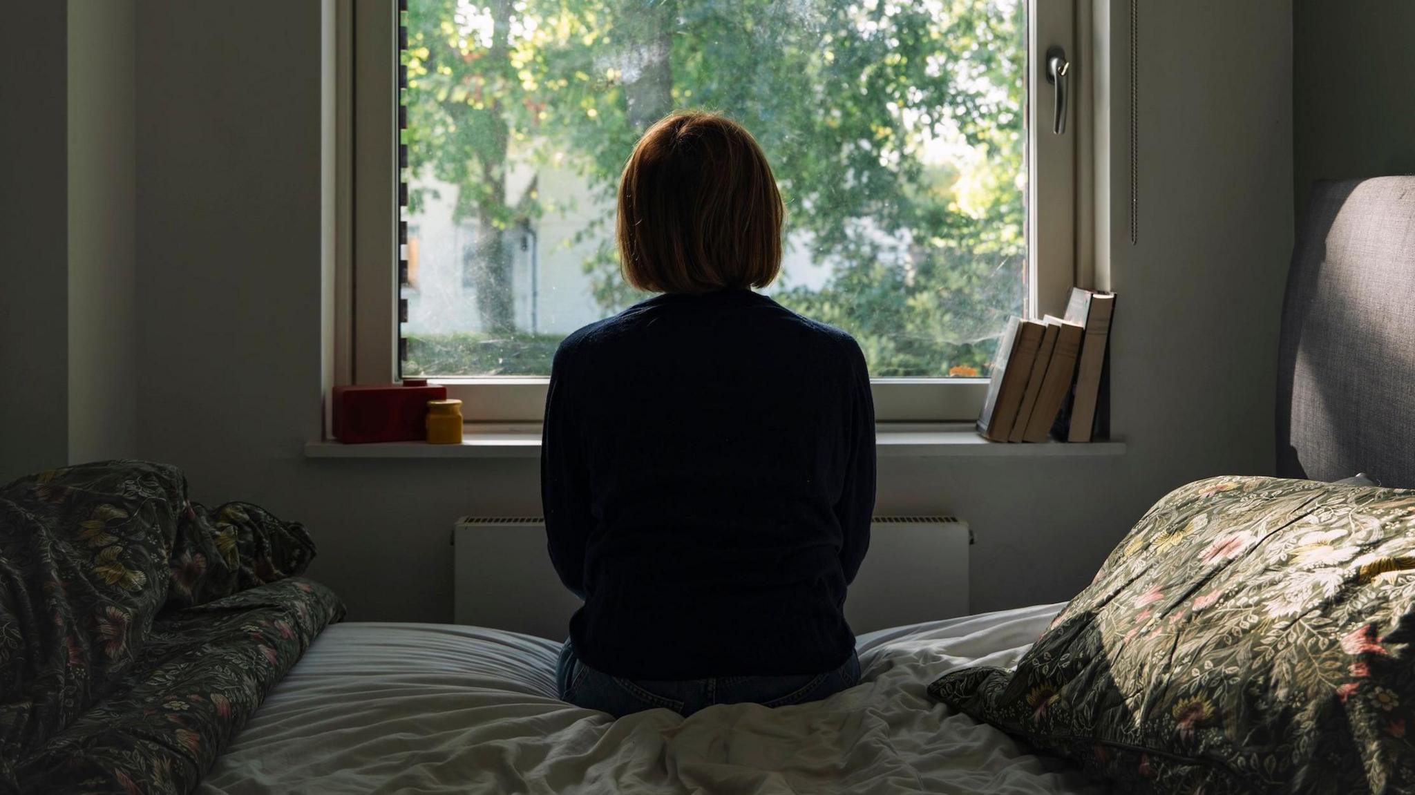 A woman looks out of a window while sitting on a bed.