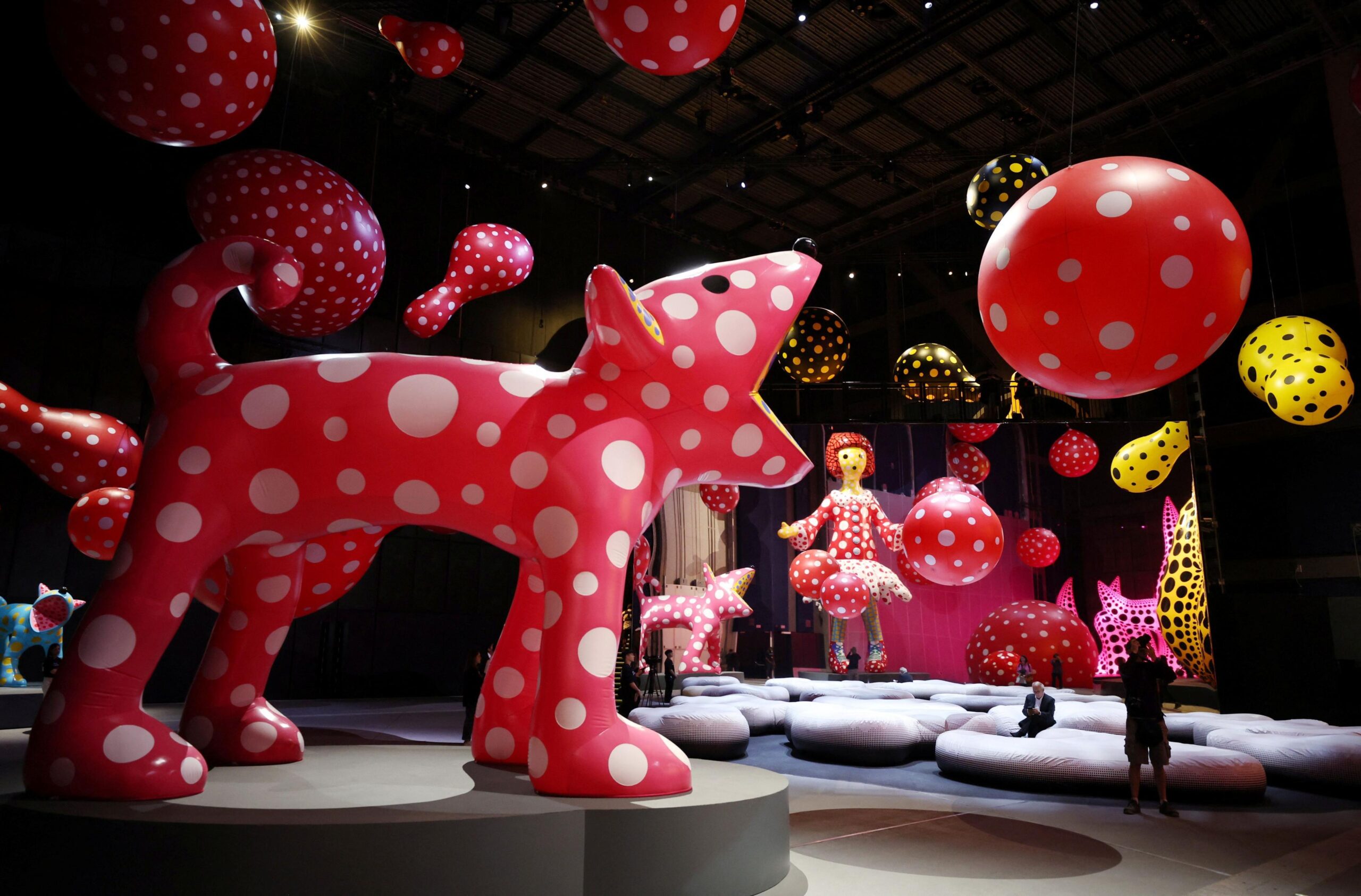 A series of large colourful polka dot sculptures in a dark gallery including a cat, a girl and balls