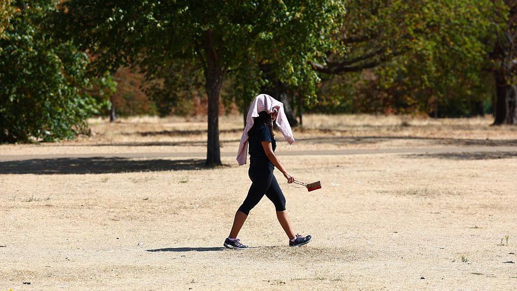 Person walking across a very dry and brown park with a towel over their head, providing shelter from the hot weather