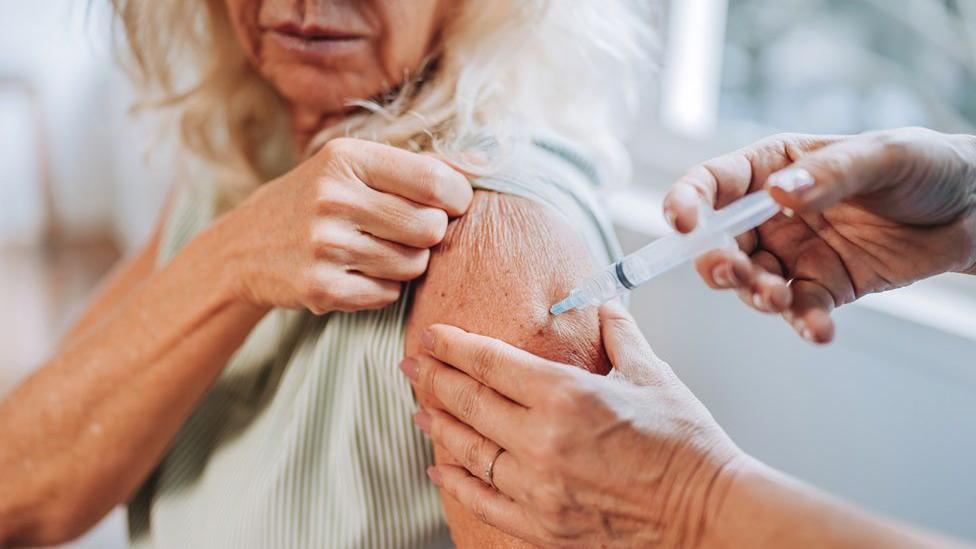 Close-up of a person receiving an injection in the upper arm, with a healthcare worker holding a syringe and supporting the arm. The image focuses on the vaccination procedure in a clinical setting, with the background softly blurred.