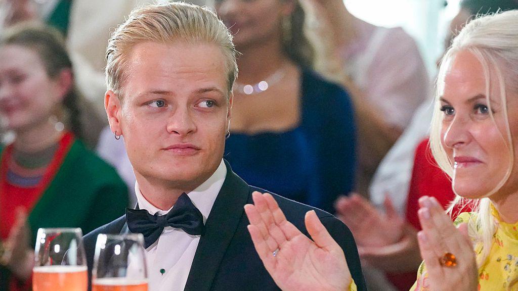 A young man with blond hair and a bow tie looks on as a woman in a yellow dress claps