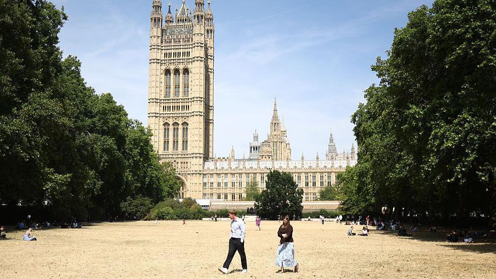 People walk on sun scorched grass at Victoria Tower Gardens, backdropped by the Houses Of Parliament in central London