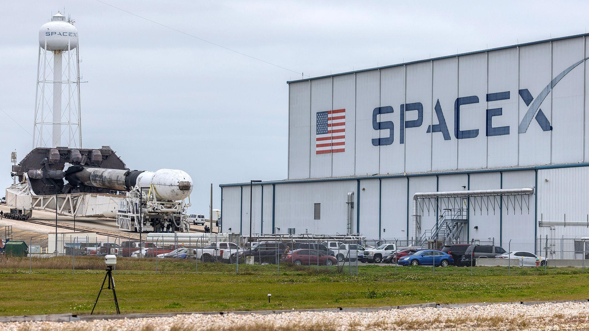 A view of a warehouse with the US flag and the word 'SpaceX' on the side with the Falcon 9 rocket in the background against a cloudy sky at the Kennedy Space Center in Cape Canaveral, Florida, as it is moved a day before launch on 14 January 2026.