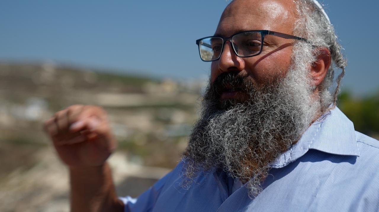 Yehuda Shimon, a large, bearded man wearing glasses, gestures with his hand