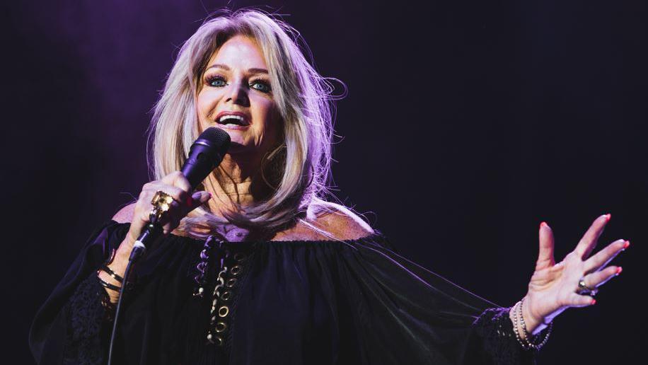 Bonnie Tyler on stage singing. She wears a black long sleeve dress and holds the microphone in one hand, with the other outstretched. Her long blonde hair is loose.