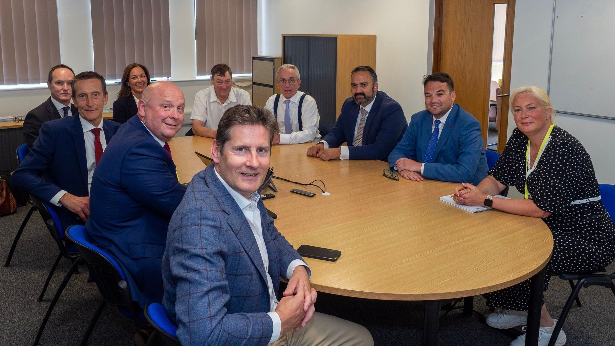 A group of eight men and two women sit around a round wooden table in a small office meeting room. They are wearing formal business attire and are facing the camera.