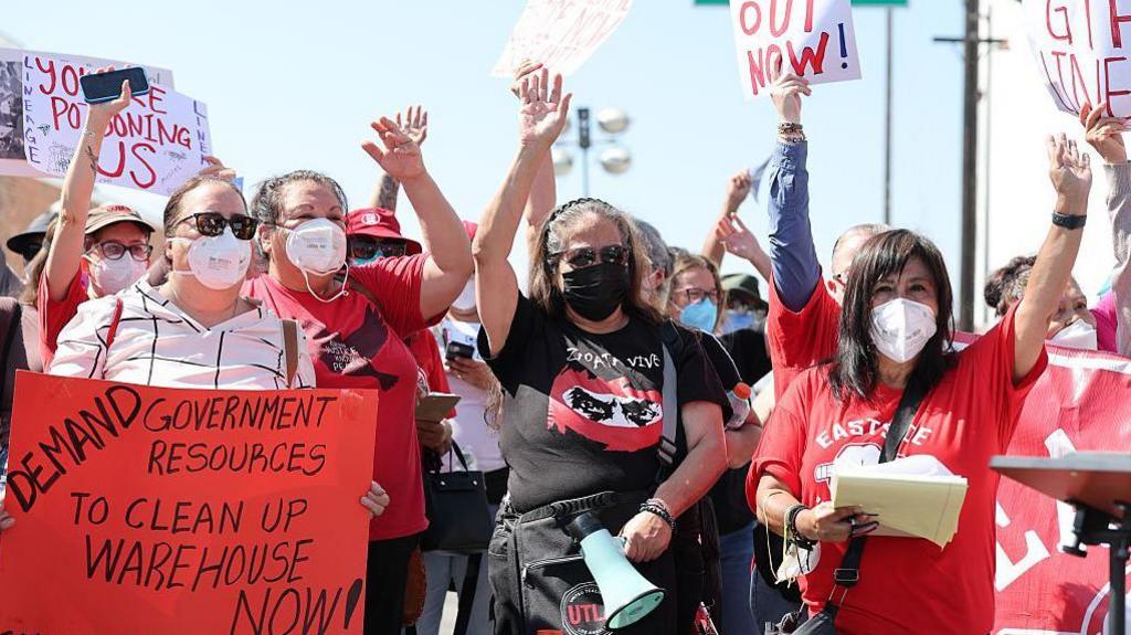 Residents in red and white and black shirts carry signs and wear masks and raise their hands during a protest. One sign reads: "Demand government resources to clean up warehouse now!"