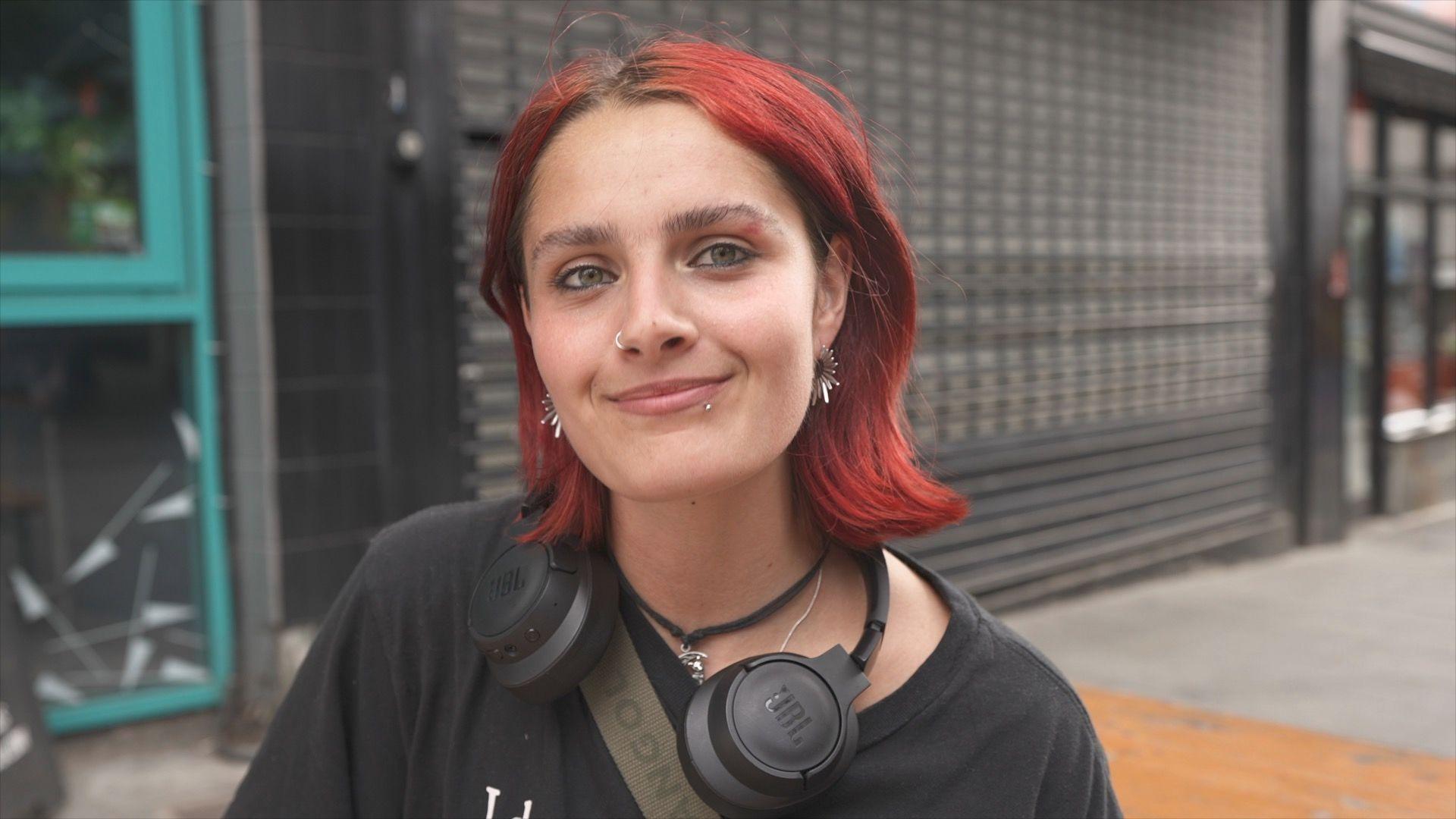 young woman with short red hair in the foreground, wearing a black T-shirt, black over-ear headphones around the neck, a cross-body strap, a nose ring and earrings. The person is facing the camera on a pavement outside a building. Behind the person is a dark tiled wall and metal security shutter, with a turquoise-framed doorway visible on the left and part of a street scene on the right. No bags, bins or markers are visible.