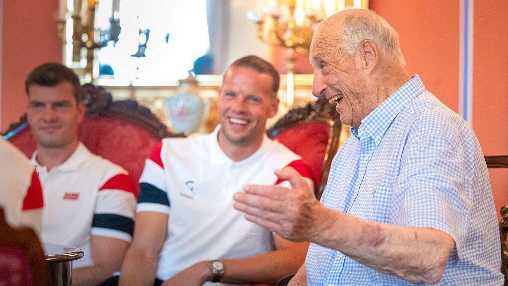 The king wearing a checkered collared t-shirt laughs as he talks. Two footballers seated on a sofa laugh in the background.