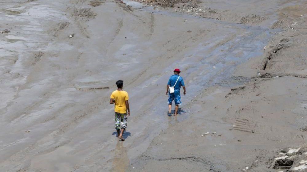 Two men walk through thick mud and water in a barren landscape