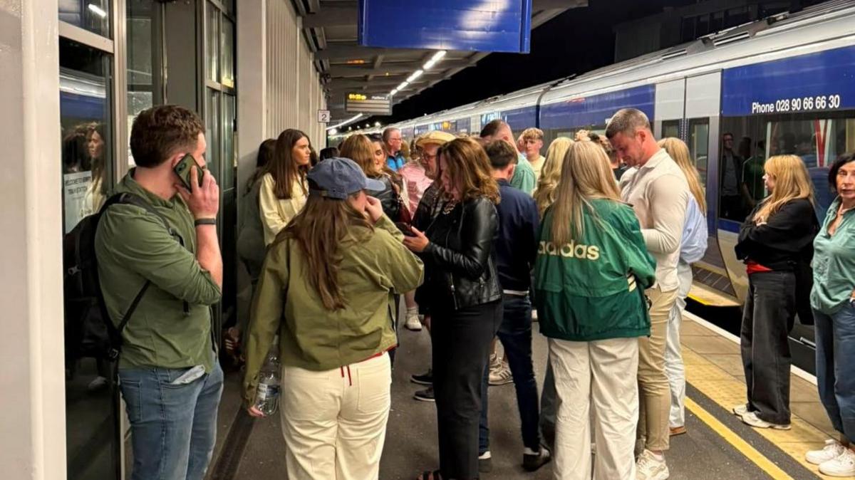 A group of passengers standing on a train platform at night.  Many are looking at their mobile phones
