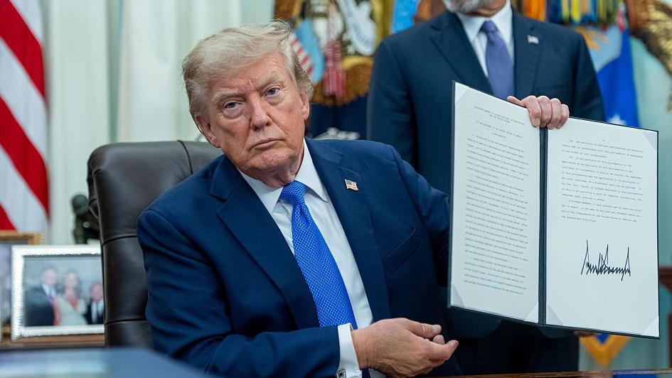 Trump, wearing a blue suit and sitting at desk in the Oval Office, holding up a signed executive order