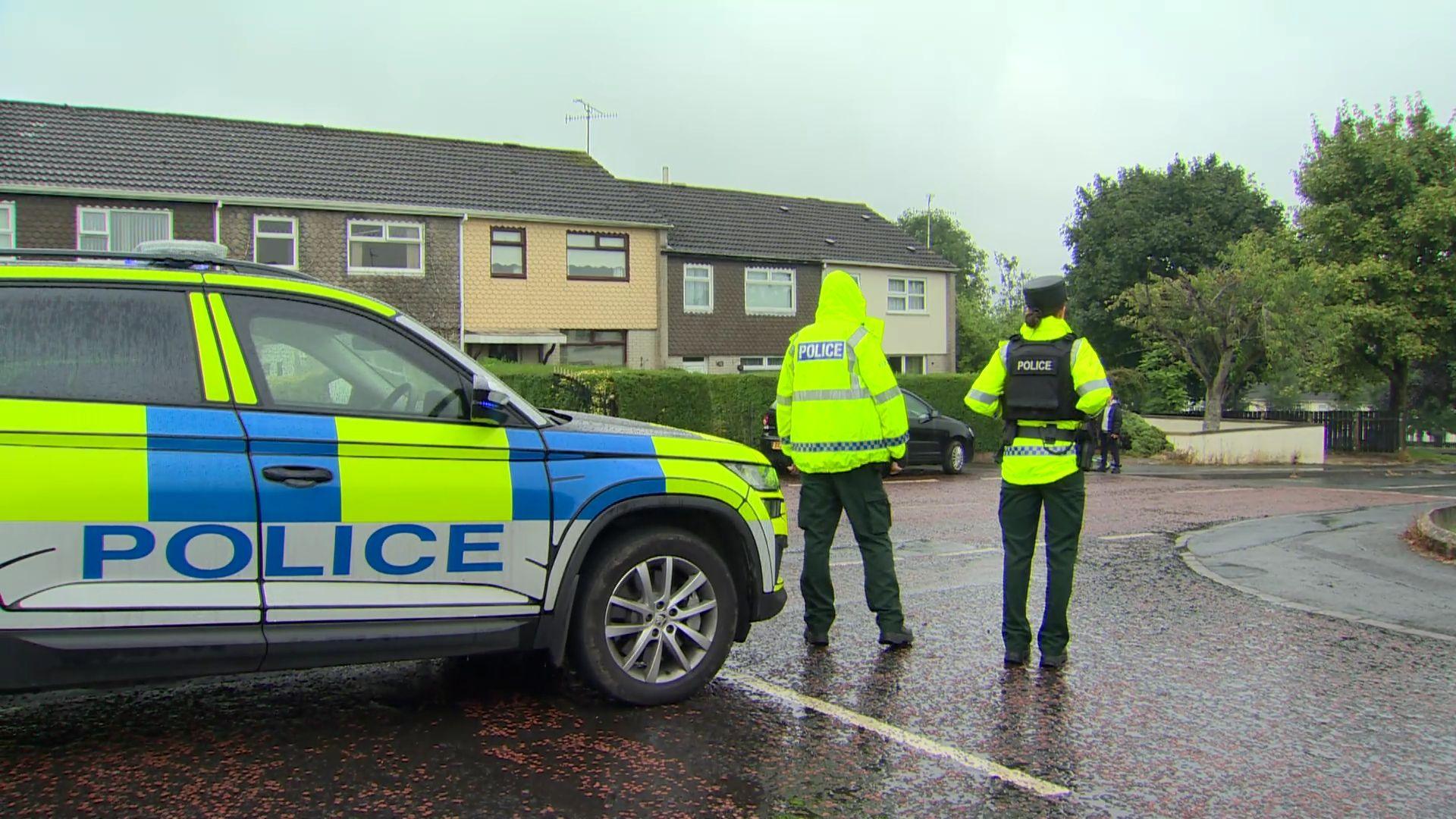 Two police officers stand beside a car, wearing hi-vis waterproof coats and police vests.
