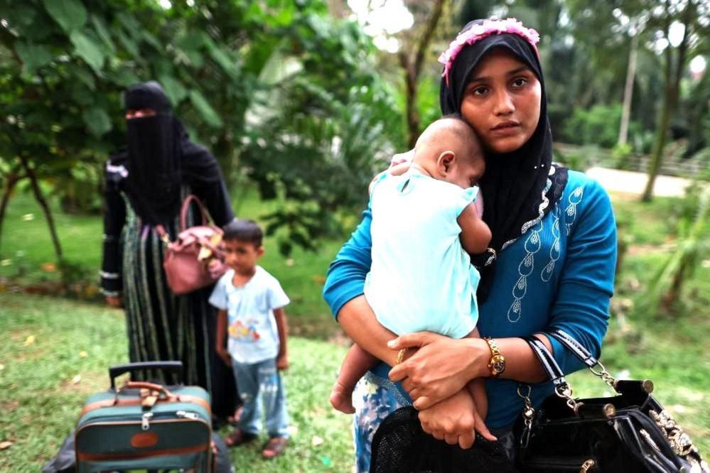A Rohingya woman carrying a baby is pictured in the foreground while another lady stands with a young boy and a suitcase in the background