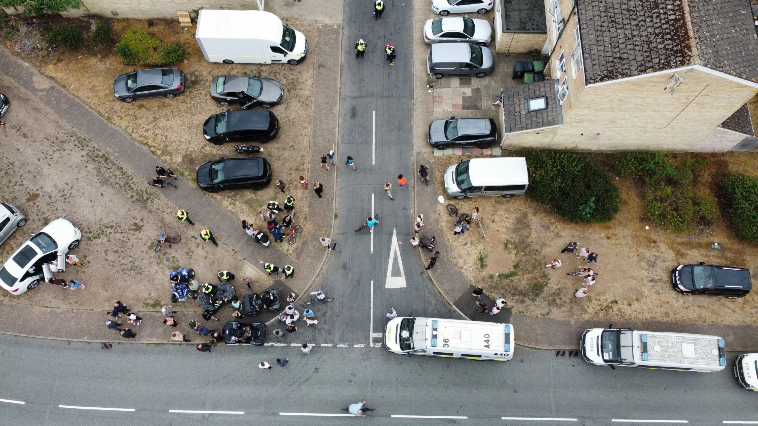 An aerial photo shows people gathered on a street corner with others elsewhere on the road. There are a number of vehicles parked on the street and in driveways.
