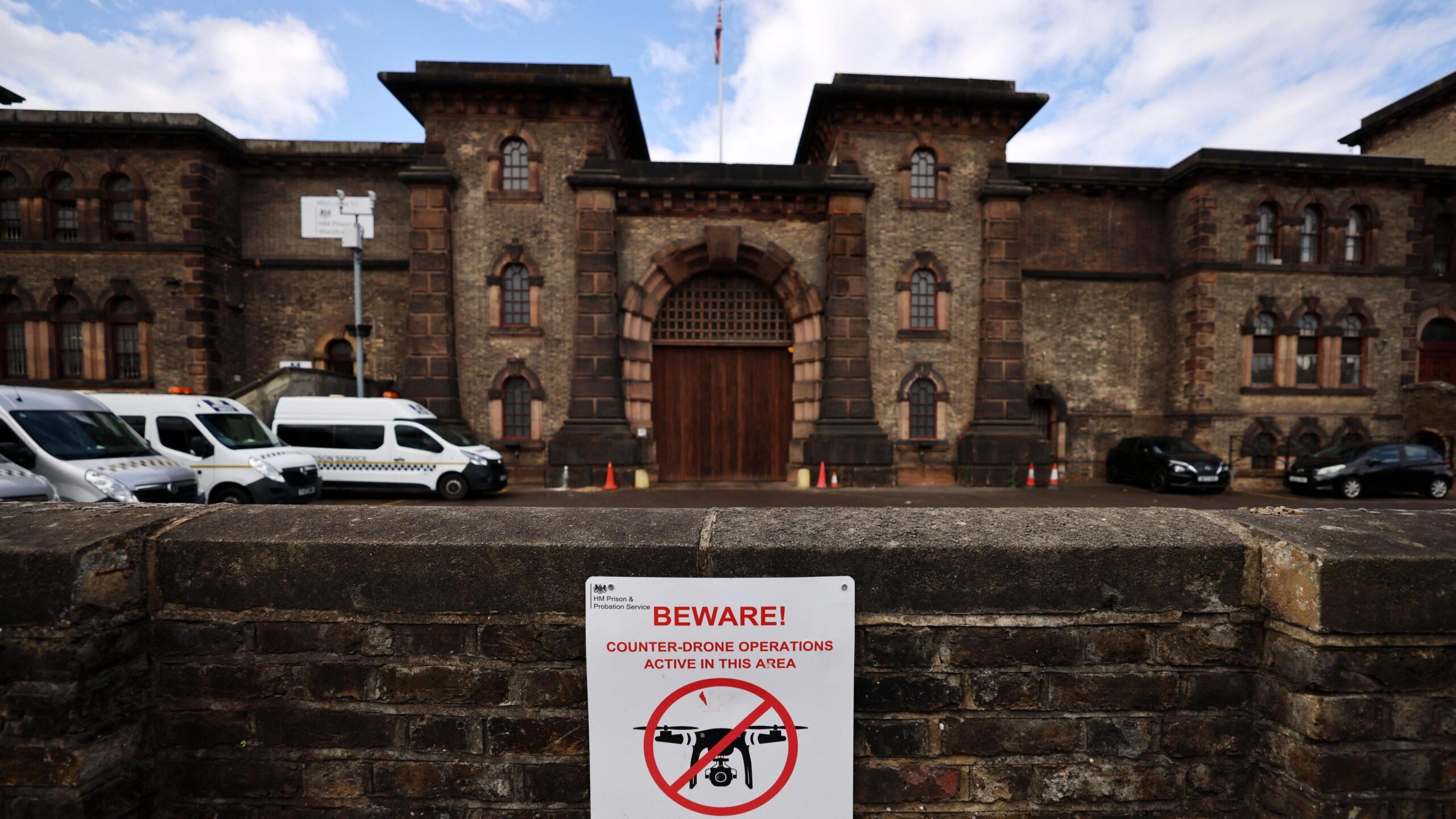 The entrance to HMP Wandsworth, a stone building resembling a castle, with a low wall in front of it bearing a sign saying "Beware counter-drone operations active in this area" with a drawing of a drone with a red circle around it and a diagonal red line through it