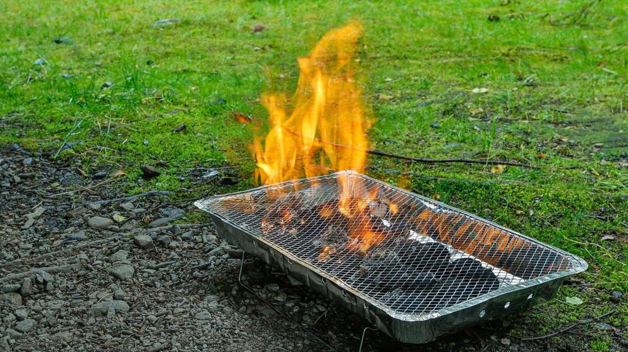 A disposable barbecue straddles a soily patch and grass as it burns.