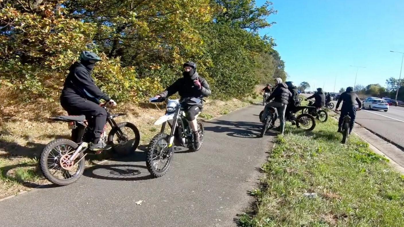 A group of riders mostly dressed in black, some with helmets and face coverings. They are on a grass verge and path next to a busy road, but appear to be stopped and are facing in different directions.