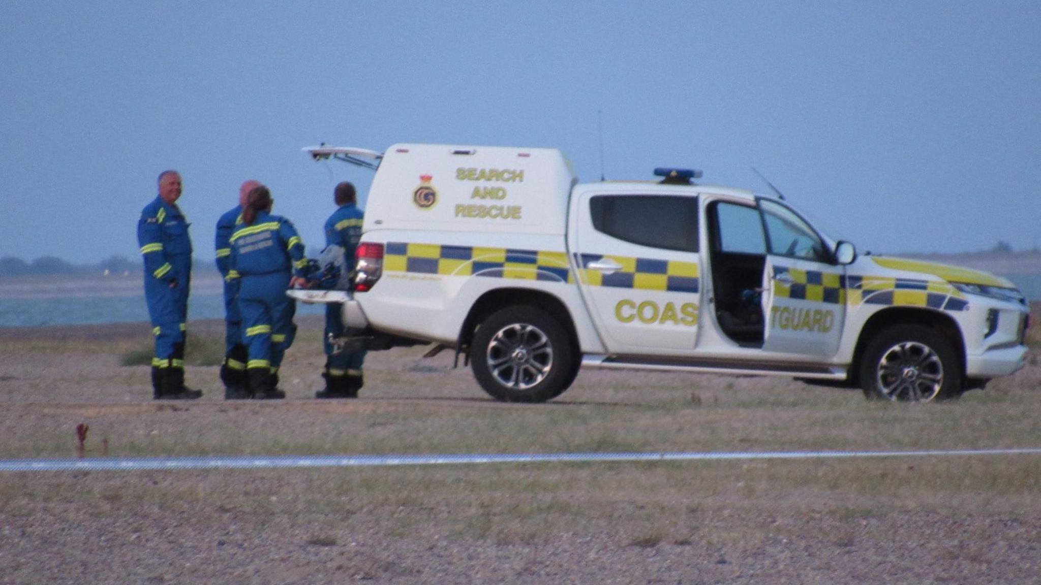 A white Coastguard vehicle with the words "Search and Rescue" and "Coastguard" printed on the side. It is on a beach and beyond it is sea. There are four people in blue boiler suits at its rear door, which is open.
