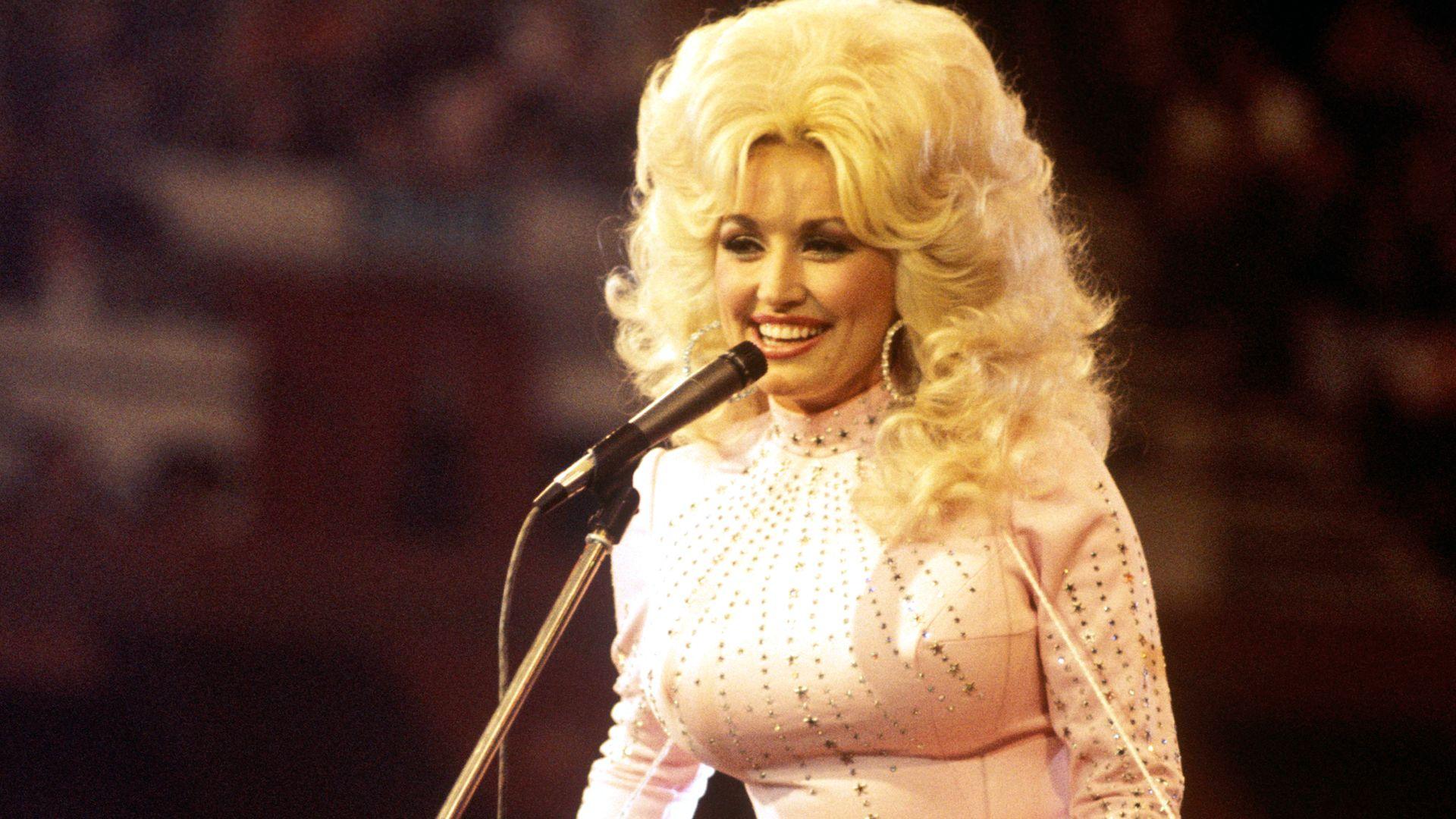 Dolly Parton smiles on stage wearing a white rhinestone jumpsuit.