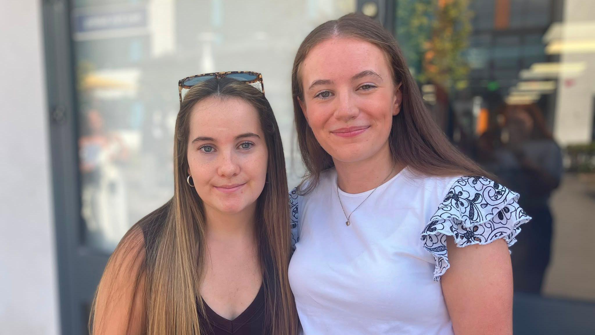 Beatrice and Amelia stand side by side outdoors, looking towards the camera. Amelia is wearing a dark sleeveless top with sunglasses resting on their head, while Beatrice is wearing a white top with patterned ruffled sleeves. The background is softly blurred and shows a modern building entrance.