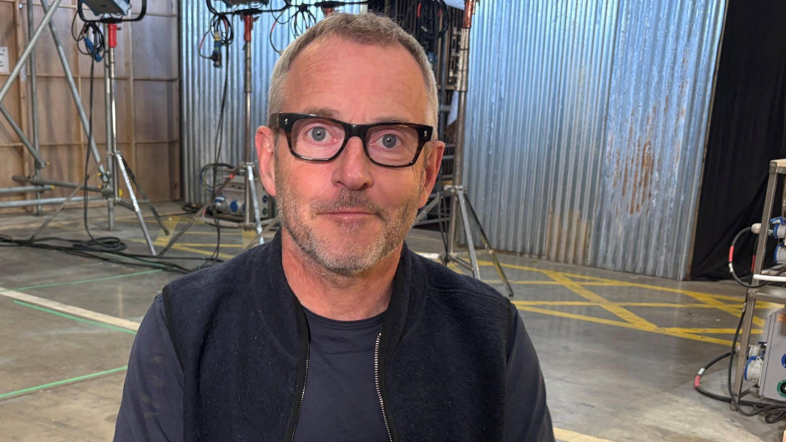 Rob Bullock sitting in the studio. He has short, brown hair and is wearing black-rimmed glasses and a black top. Corrugated iron walls and metal lights can be seen in the background.