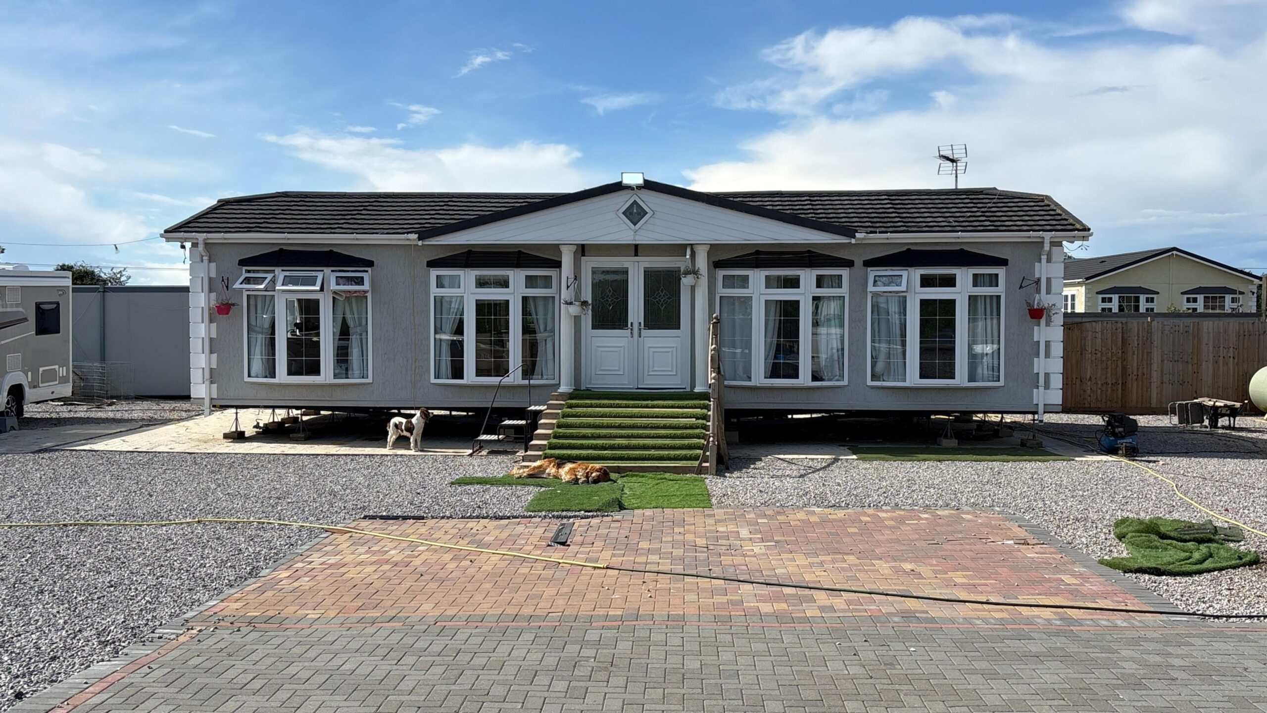 A wide shot of a static caravan. It is set back from where the camera is stationed, with an expanse of block paving and a stone driveway in the foreground. A small stairway leads up to the caravan's double, uPVC-type doors. Either side of the doors are two sets of windows, meaning across the front there are four sets of windows in total. The caravan is symmetrical and imposing against a light blue, partly cloudy sky.